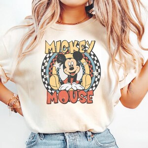 May include: Cream-colored t-shirt featuring a retro design of Mickey Mouse. The graphic includes Mickey in a seated pose, surrounded by a circular design with the words "Mickey Mouse" in a vintage font. The design incorporates a checkered pattern.