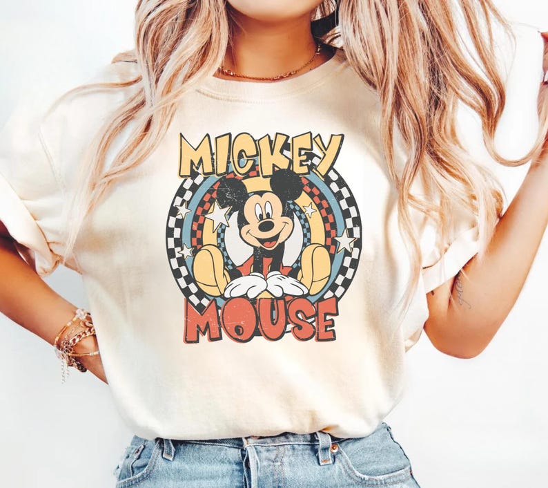 May include: Cream-colored t-shirt featuring a retro design of Mickey Mouse. The graphic includes Mickey in a seated pose, surrounded by a circular design with the words "Mickey Mouse" in a vintage font. The design incorporates a checkered pattern.