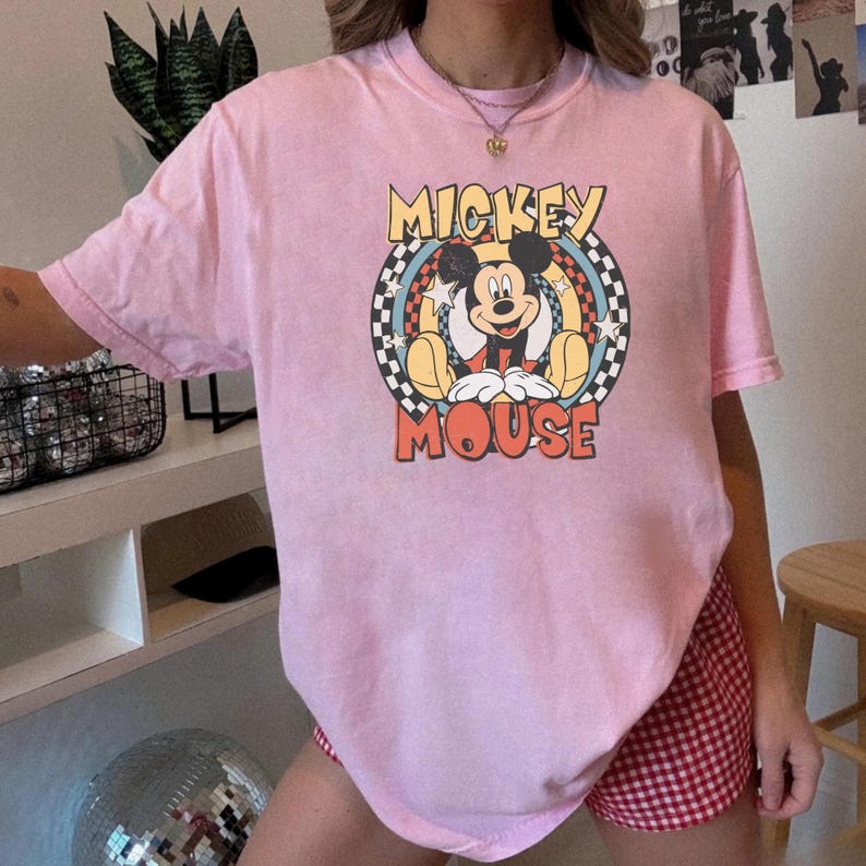 May include: A light pink t-shirt featuring a graphic of Mickey Mouse. The design includes the words "Mickey Mouse" in a retro font, with Mickey in the center. The shirt has short sleeves.