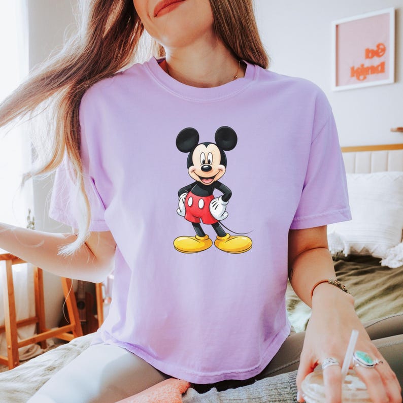 May include: A light purple short-sleeve t-shirt featuring a centered graphic of Mickey Mouse. The character is depicted with his signature black ears, red shorts, white gloves, and large yellow shoes, standing with hands on his hips.