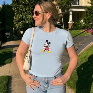 Puede incluir: Una camiseta de manga corta en color azul claro con un gr&aacute;fico central de Mickey Mouse vistiendo pantalones cortos rojos y zapatos amarillos. La prenda se combina con vaqueros azules y un peque&ntilde;o bolso de hombro color crema con correa fina.