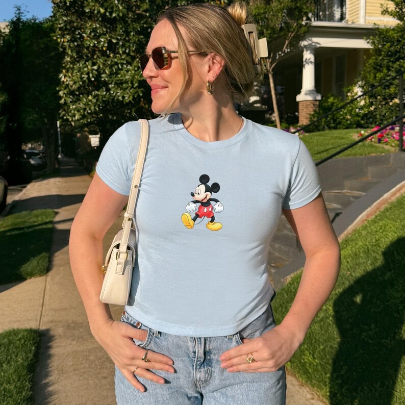 Puede incluir: Una camiseta de manga corta en color azul claro con un gr&aacute;fico central de Mickey Mouse vistiendo pantalones cortos rojos y zapatos amarillos. La prenda se combina con vaqueros azules y un peque&ntilde;o bolso de hombro color crema con correa fina.