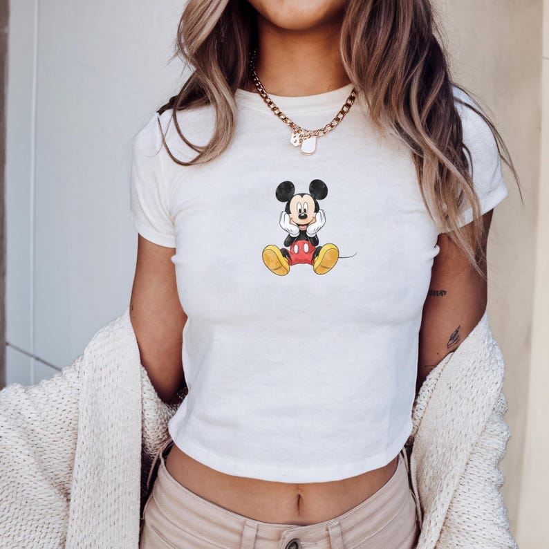 May include: White crop top with a seated Mickey Mouse design. The cartoon character is in black, red, and yellow. The top has short sleeves and a crew neck. The shirt is a casual style.