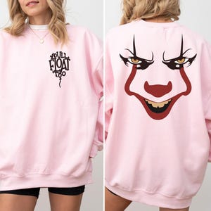 May include: A light pink crewneck sweatshirt featuring a black graphic on the front left chest that reads "YOU'LL FLOAT TOO" inside a balloon shape. The back of the sweatshirt displays a large, detailed graphic of a clown face with red makeup.