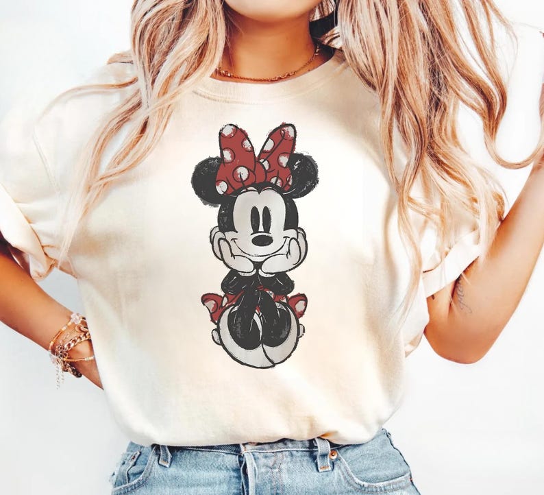 May include: Cream-coloured t-shirt with a distressed graphic of Minnie Mouse. Minnie is shown with a red polka-dot bow, black ears, and a red skirt. The design has a vintage, worn appearance.