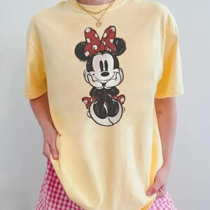 May include: A pale yellow t-shirt with a Minnie Mouse graphic. The graphic depicts Minnie Mouse in a seated pose, with a red polka dot bow and matching details. The shirt has a crew neck and short sleeves.