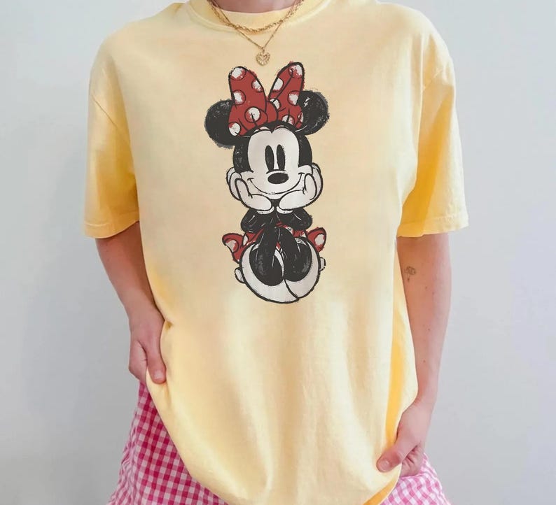 May include: A pale yellow t-shirt with a Minnie Mouse graphic. The graphic depicts Minnie Mouse in a seated pose, with a red polka dot bow and matching details. The shirt has a crew neck and short sleeves.