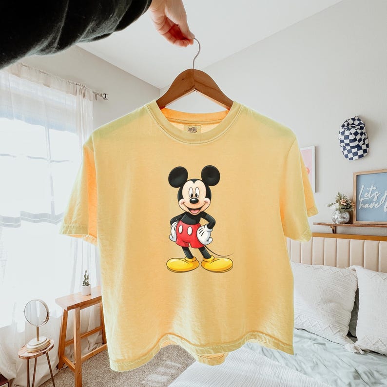 May include: A pale yellow short-sleeve t-shirt featuring a centered graphic of Mickey Mouse. The character is depicted with his signature black ears, red shorts, white gloves, and yellow shoes, standing with hands on his hips against the solid yellow fabric.