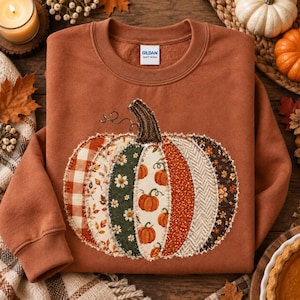 May include: A rust-coloured crewneck sweatshirt featuring a large, central pumpkin appliqué. The pumpkin is constructed from various fabric patches with plaid, floral, and herringbone patterns in shades of orange, green, and cream. A Gildan tag is visible.