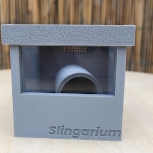 Può includere: Un contenitore grigio stampato in 3D con un coperchio testurizzato e un pannello frontale trasparente. La parola Slingarium è impressa sulla parte anteriore inferiore. All'interno della scatola è visibile un tubo cilindrico grigio come rifugio.