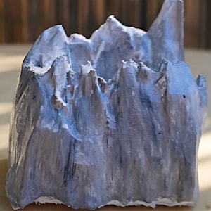 May include: A small, hollow, mountain-shaped container with a jagged, uneven top edge. The exterior surface is painted in shades of grey and white, featuring a textured, layered finish that mimics the appearance of rugged, rocky terrain.
