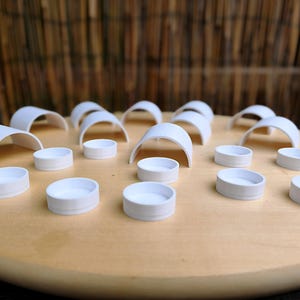 Könnte beinhalten: Ein Set aus weißen Kunststoff-Spielfiguren, die auf einem runden Holzbrett angeordnet sind. Die Sammlung umfasst mehrere bogenförmige, brückenartige Strukturen sowie zahlreiche kleine, kreisförmige und flache Behälter oder Spielsteine.