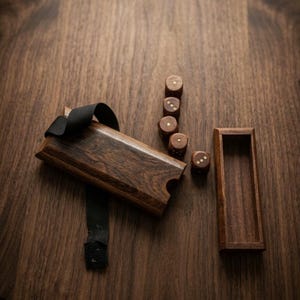 May include: A dark brown wooden dice box with a black fabric strap and a matching open rectangular wooden tray. Four small wooden dice with light-colored pips are arranged in a line next to the box. The text ACHCHHA SA BACKGROUND is visible.