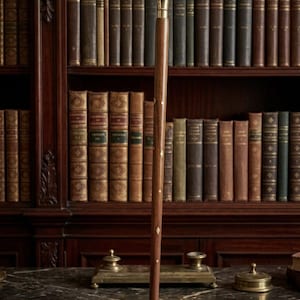 May include: A walking stick crafted from dark brown wood featuring a polished gold-tone metal handle and small, diamond-shaped brass inlays spaced along the length of the shaft. The stick terminates in a matching gold-tone metal tip at the base.