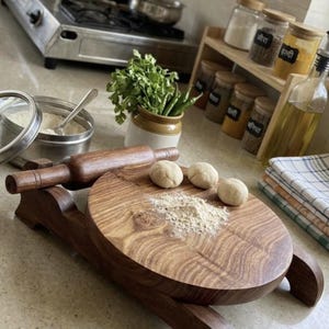 May include: A round wooden rolling board with a matching wooden rolling pin resting on a stand. Three small balls of dough and a dusting of flour are on the board. A metal container with flour, a small ceramic pot with herbs, and spice jars are nearby.