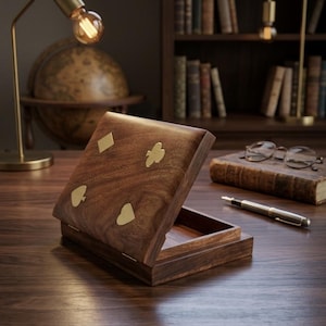 Peut inclure: Une boîte rectangulaire en bois avec un couvercle à charnière orné de quatre symboles de cartes à jouer en laiton incrusté : un carreau, un trèfle, un pique et un cœur. La boîte est fabriquée en bois brun foncé avec un grain visible et poli.