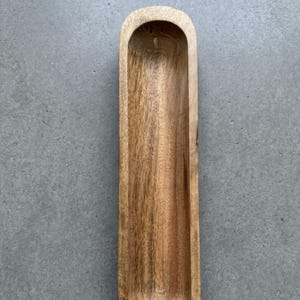 Op de afbeelding: Een lang, smal, rechthoekig houten dienblad met afgeronde uiteinden en een diep, glad, concaaf midden. Het dienblad heeft een natuurlijk lichtbruin houtnerfpatroon met zichtbare variaties in kleur en textuur over het gehele oppervlak.
