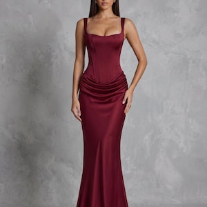 Puede incluir: Un vestido largo de color burdeos con un corpiño estructurado estilo corsé, tirantes anchos y escote cuadrado. La falda está confeccionada en un tejido satinado liso con detalles fruncidos en la cintura, cayendo hasta el suelo.