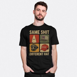 Puede incluir: Una camiseta negra de manga corta con un estampado gráfico que incluye el texto SAME SHIT DIFFERENT HAT. El diseño presenta cuatro iconos cuadrados que muestran una capucha blanca, una gorra con bandera confederada, una gorra militar y una gorra roja.