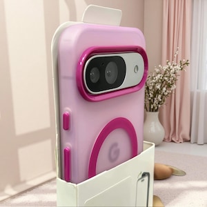 Può includere: Una custodia per smartphone rosa chiaro con una cornice per fotocamera rosa scuro e un accento circolare rosa scuro abbinato sul retro. La custodia presenta un modulo fotocamera argentato con due lenti e un flash, oltre a pulsanti laterali rosa scuro.
