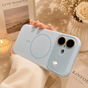 Op de afbeelding: Een lichtblauwe smartphonehoes met een cirkelvormige magnetische oplaadring aan de achterzijde en een glanzende, verhoogde cameralensbeschermer met goudkleurige randen rondom twee zwarte lenzen en een kleine ronde flitsopening.