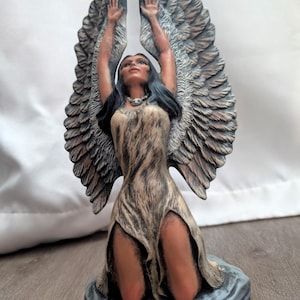 May include: A detailed figurine of a Native American woman kneeling with her arms raised. She wears a fringed dress and a necklace. Large, detailed wings extend upwards behind her. The base is a textured, grey stone-like material.