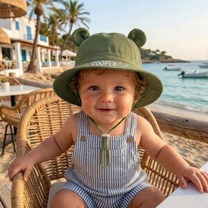 Peut inclure: Un bébé souriant porte un chapeau vert avec des oreilles d'ours et le nom "COOPER" brodé dessus. Le bébé porte également une salopette à rayures bleues et blanches. Le bébé est assis dans une chaise en osier près de la plage.