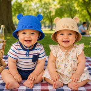 May include: Two smiling babies wearing matching sun hats with bear ears. One hat is blue with the name "Ethan" embroidered on it, and the other is tan with "Mia" embroidered. They are sitting on a picnic blanket.