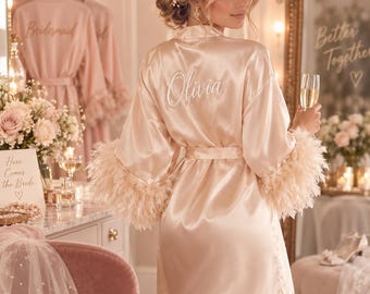 May include: A champagne-colored satin robe featuring long sleeves with feathered cuffs and a tie waist. The back of the robe is embroidered with the name Olivia in white script lettering. The fabric has a smooth, reflective finish.