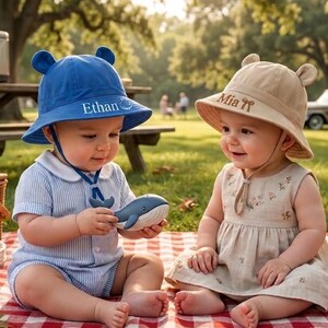 May include: Two babies wearing blue and tan sun hats with bear ears and the names "Ethan" and "Mia" embroidered on them. One baby holds a blue whale toy. They are sitting on a red and white checkered blanket.