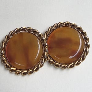 Könnte beinhalten: Ein Paar runde Clip-Ohrringe mit glatten, durchscheinenden bernsteinfarbenen Cabochons, die in polierte goldfarbene Metallfassungen eingefasst sind. Jeder kreisförmige Ohrring ist von einem dekorativen gedrehten Seilrand umgeben, was ein symmetrisches Design ergibt.