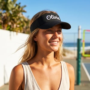 May include: Black visor with the name "Olivia" embroidered in white script. The visor has a curved brim. The background includes a beach and a volleyball net. The person wearing the visor has blonde hair.