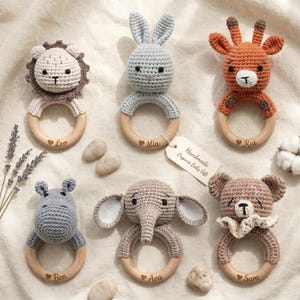 May include: Six crochet animal rattles on wooden rings, featuring a lion, rabbit, giraffe, hippo, elephant, and bear. Each ring is engraved with a name and a heart symbol. A paper tag attached to the elephant rattle reads Handmade Organic Baby Gift.