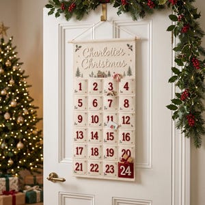Peut inclure: Un calendrier de l'Avent en tissu crème suspendu à une porte blanche, avec le texte "Charlotte's Christmas" en haut. Il comporte vingt-quatre poches carrées avec des chiffres rouges brodés, des illustrations d'arbres verts et du houx.