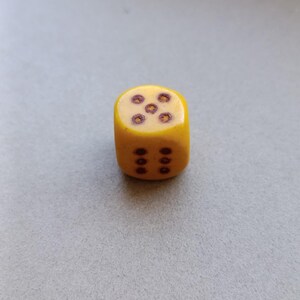 Può includere: Un dado a sei facce di colore giallo con angoli arrotondati. La faccia superiore presenta cinque punti circolari marrone scuro disposti a quinconce, mentre la faccia anteriore mostra sei punti marrone scuro in due colonne verticali da tre.