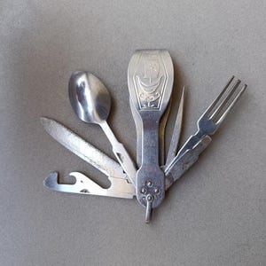 May include: A silver-toned multi-tool camping utensil set featuring a folding spoon, fork, knife, and bottle opener. The central handle displays an engraved sailboat design. The metallic components are arranged in a fan shape, showcasing various integrated dining and utility implements for outdoor use.