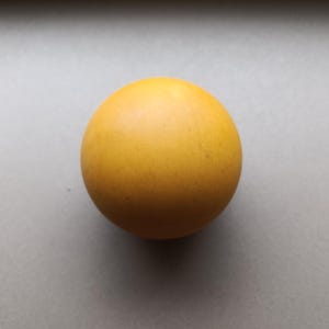 Op de afbeelding: Een solide, glad, bolvormig object in een levendige mosterdgele kleur. Het oppervlak van de bal ziet er mat en uniform uit, met een consistente textuur over het geheel. Het is een eenvoudige, ronde, driedimensionale vorm zonder zichtbare markeringen.