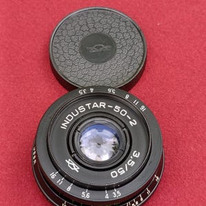 May include: Black Industar-50-2 camera lens with a matching lens cap. The lens has the text "INDUSTAR-50-2" and "3.5/50" printed on it. The lens cap is dark green with a textured surface and a logo.