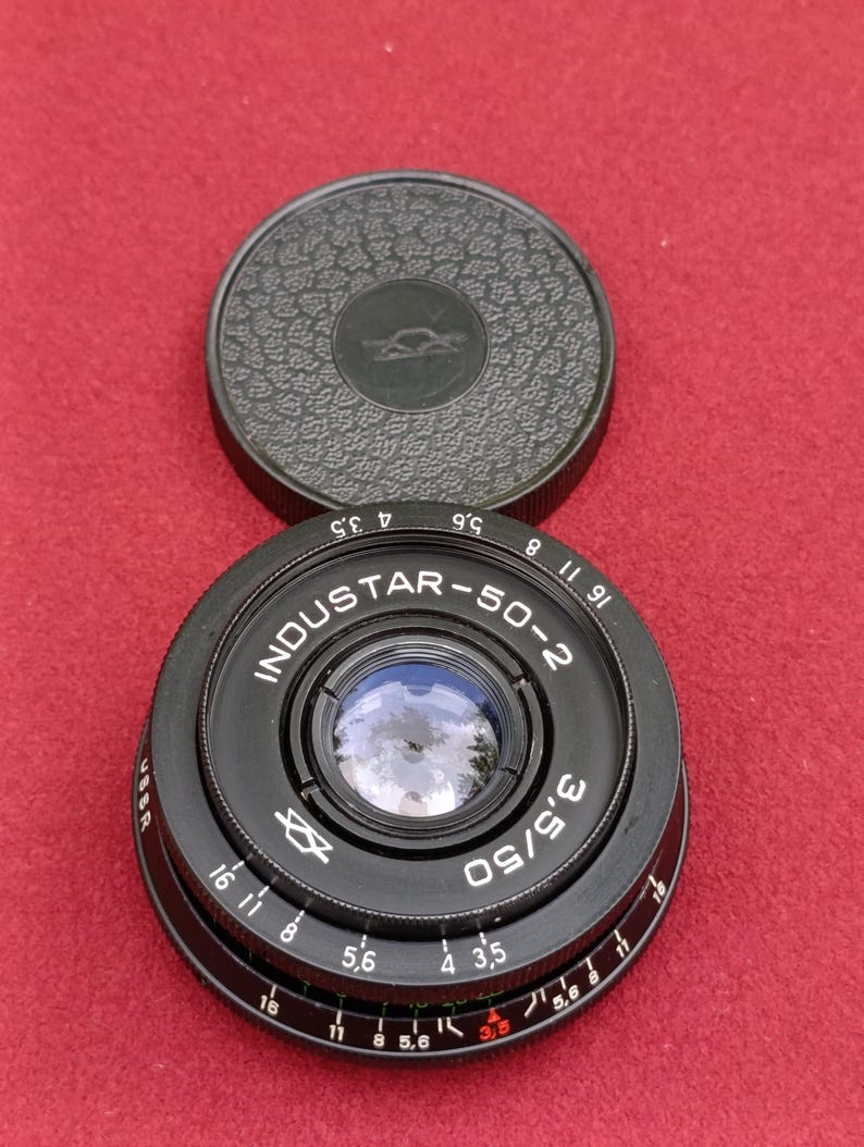 May include: Black Industar-50-2 camera lens with a matching lens cap. The lens has the text "INDUSTAR-50-2" and "3.5/50" printed on it. The lens cap is dark green with a textured surface and a logo.