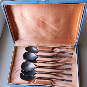 May include: A set of six silver-toned spoons with twisted handles arranged in a blue rectangular presentation box. The interior of the box is lined with peach-colored satin fabric, featuring a circular stamp on the lid's inner lining.