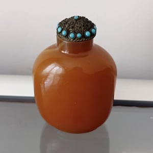 May include: A smooth, rounded, amber-coloured snuff bottle with a polished finish. The bottle features a decorative metal cap with intricate filigree detailing and a series of small, round, bright turquoise-coloured stones set around the edge.