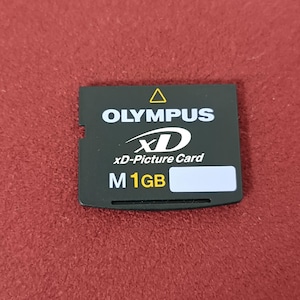 Puede incluir: Una tarjeta xD-Picture Olympus negra con texto blanco y una pegatina blanca. La tarjeta tiene el logotipo de Olympus, "xD-Picture Card" y "M 1GB". La tarjeta rectangular tiene un borde con muescas.