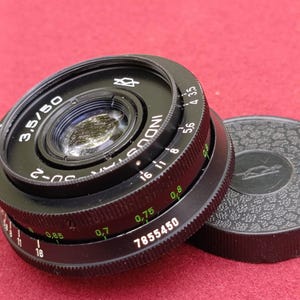 May include: A black Industar-50 3.5/50 camera lens with a matching lens cap. The lens has various markings, including f-stop numbers and the serial number 7855450. The lens cap is black with a textured surface.