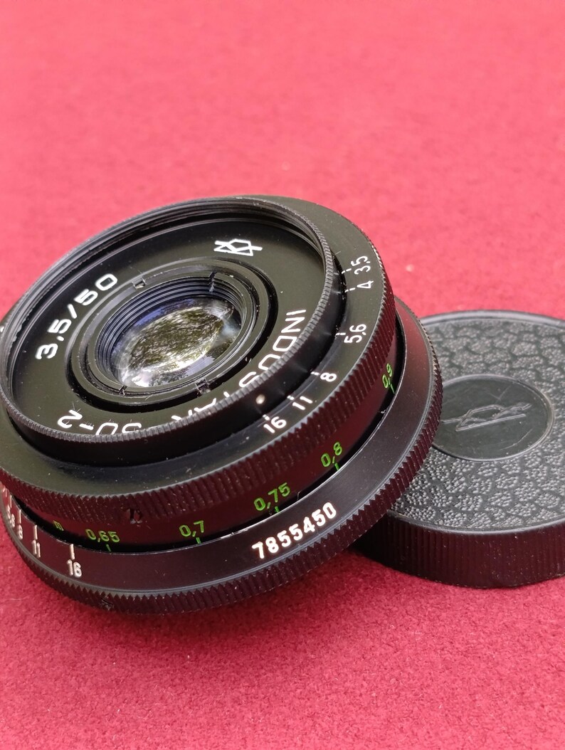 May include: A black Industar-50 3.5/50 camera lens with a matching lens cap. The lens has various markings, including f-stop numbers and the serial number 7855450. The lens cap is black with a textured surface.