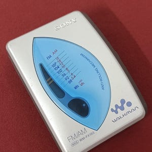 May include: A silver Sony Walkman WM-FX193 portable radio with a blue leaf-shaped display window. The device features FM/AM frequency markings, an anti-rolling mechanism label, the Walkman logo, and an AVLS indicator printed on the front surface.