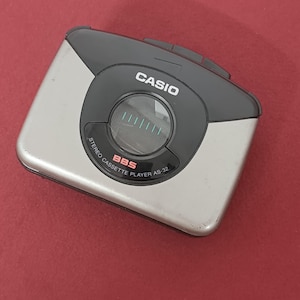 May include: A silver and black Casio stereo cassette player model AS-32. The device features a circular central window, the brand name CASIO in white text, and the red text BBS printed below the window on the black plastic control panel area.