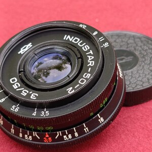 May include: Black Industar-50-2 camera lens with a black lens cap. The lens has a 50mm focal length and f/3.5 aperture. The lens has white and red markings for aperture and focus settings. The lens cap is textured.