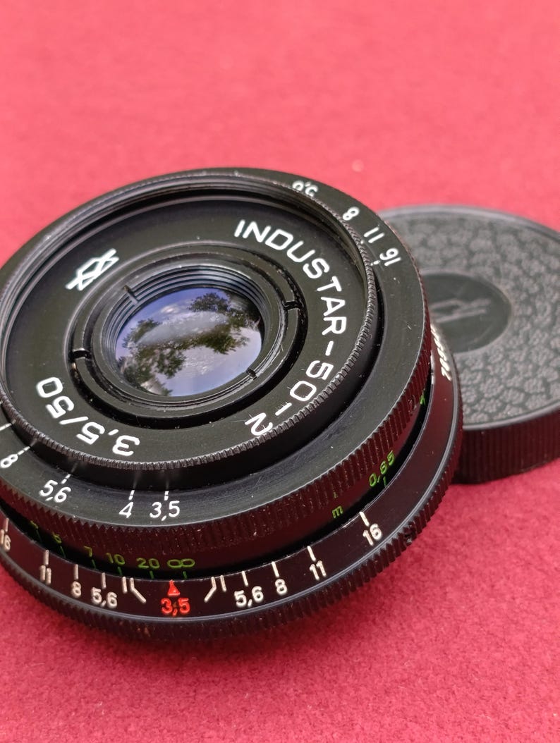 May include: Black Industar-50-2 camera lens with a black lens cap. The lens has a 50mm focal length and f/3.5 aperture. The lens has white and red markings for aperture and focus settings. The lens cap is textured.