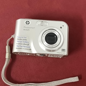 Könnte beinhalten: Silberne HP Photosmart M425 Digitalkamera mit 3x optischem Zoom. Funktionen umfassen Rote-Augen-Entfernung und In-Kamera-Effekte. Die Kamera hat 5,0 Megapixel Auflösung und eine Handschlaufe.
