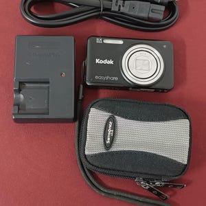 May include: A black Kodak Easyshare digital camera, charger, power cable, and a black and grey Samsonite case. The camera has a silver lens and the text "5X Wide" and "easyshare". The charger is labelled "Olympus".